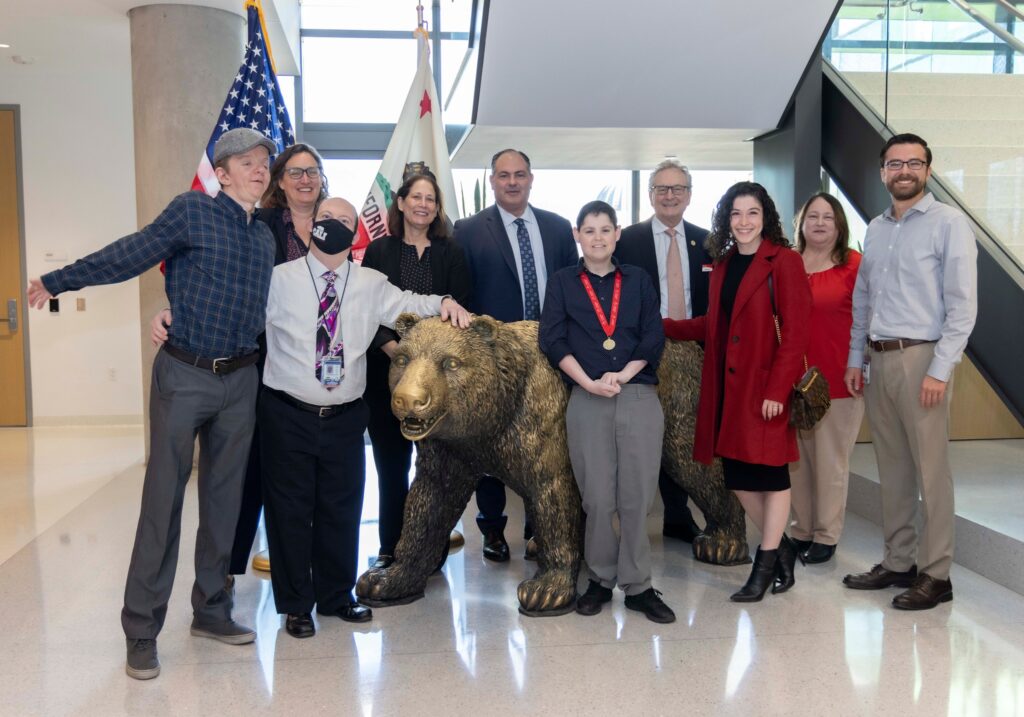 A group of Special Olympics athletes, staff, and government officials stands with a bear statue at the State Capitol building.